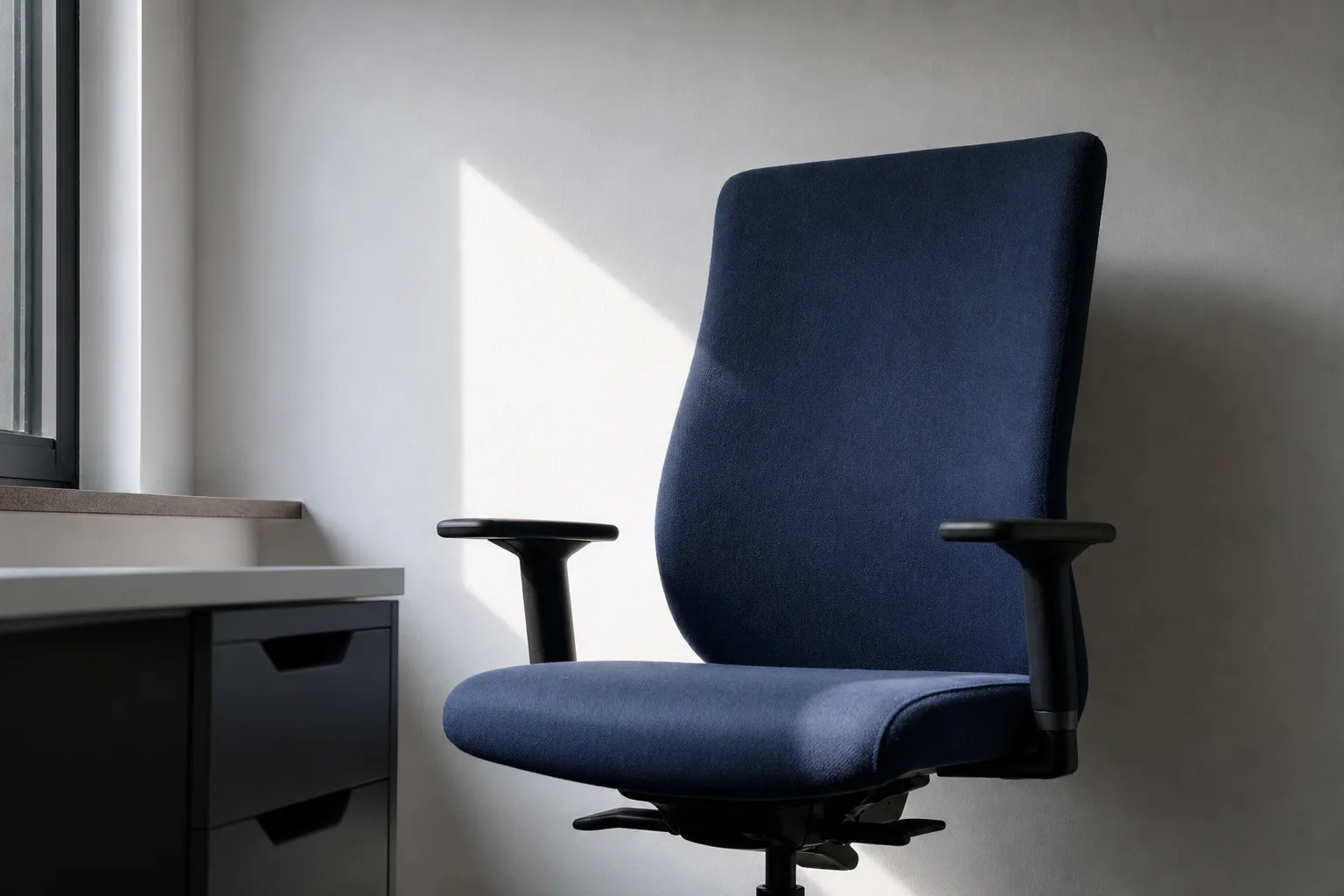 Fauteuil de bureau bleu sombre devant un mur clair, éclairé de côté par une fenêtre.
